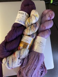 3 skeins of yarn for Love Ewe Zombie in a white with speckles, a lavender with speckles, and a solid royal purple.